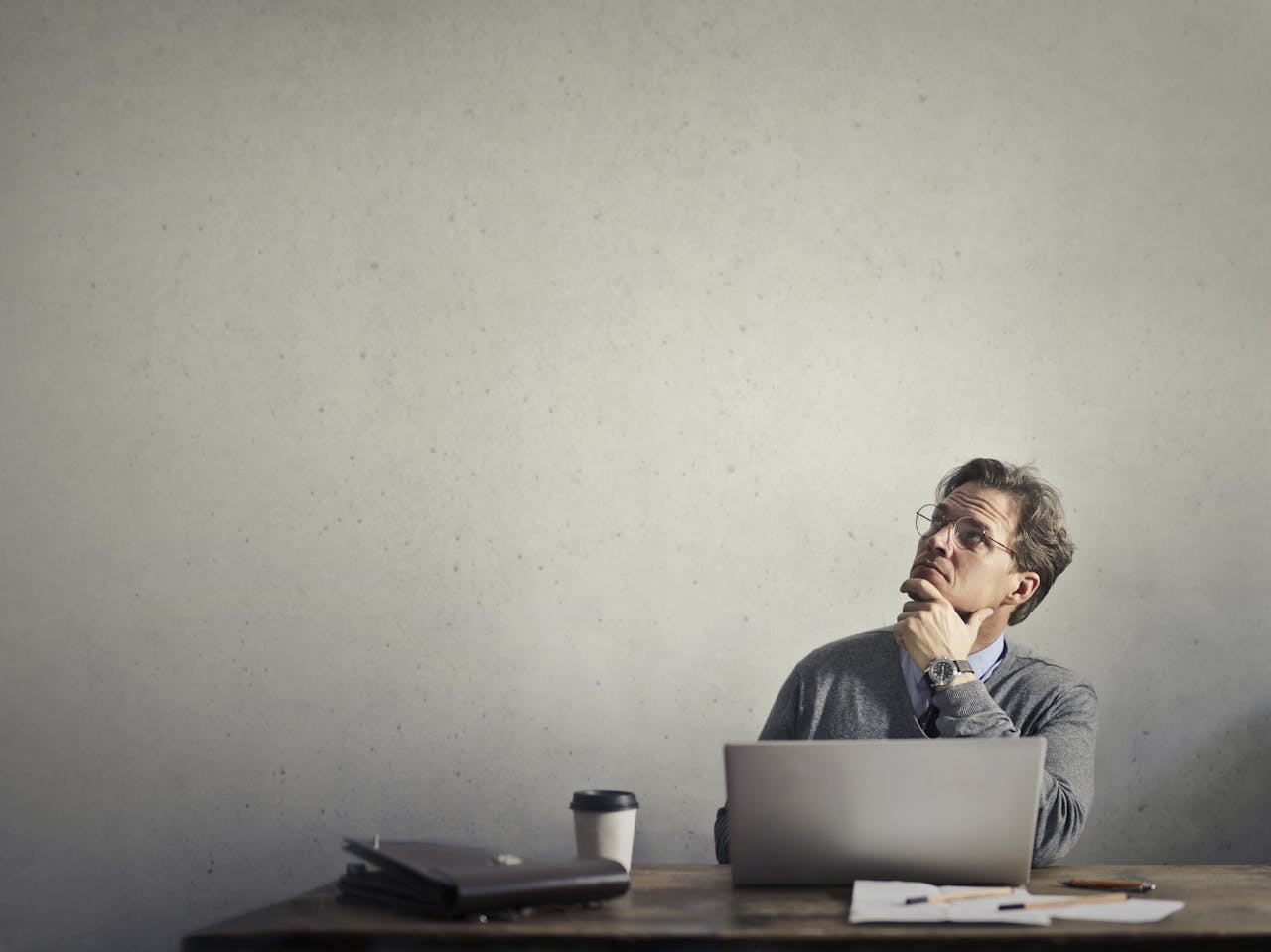 Thoughtful businessman looking up while working at his desk with a laptop and coffee.