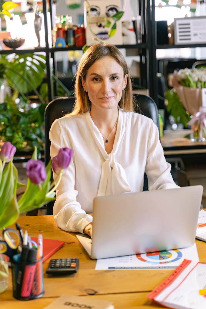 Confident woman in smart casual attire working at a vibrant, plant-filled office desk with a laptop.